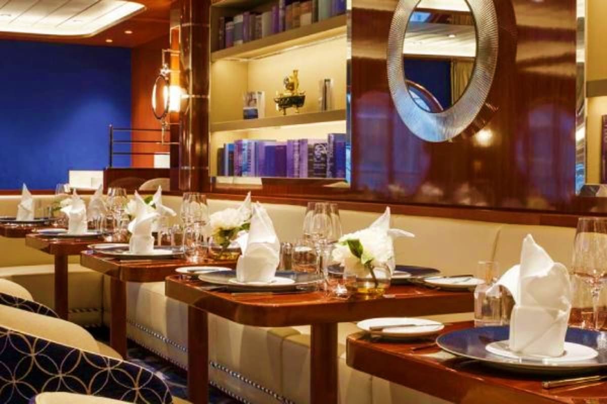 Elegant dining room with white napkins, wood tables, cream seating, illuminated shelving with books, round mirror, and blue a