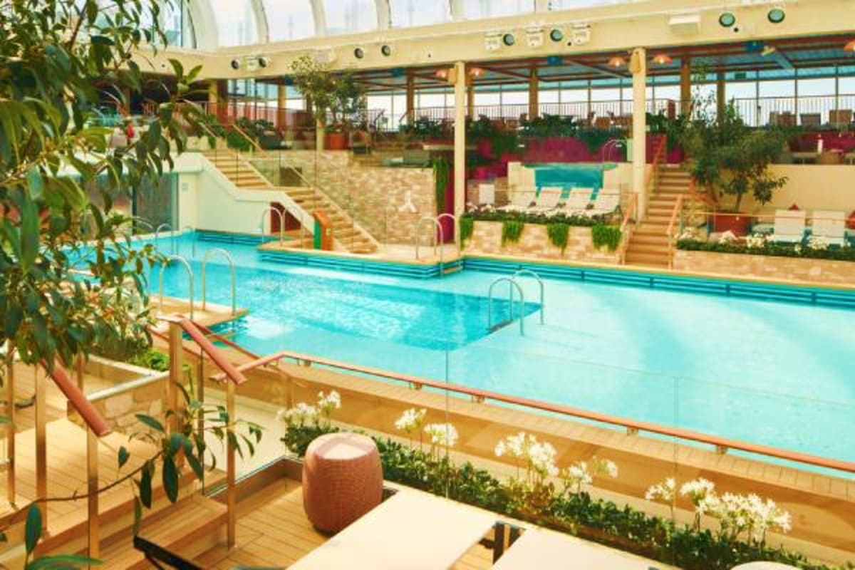 Indoor swimming pool with turquoise water, wooden deck, glass barriers, lounge chairs, potted plants, and multi-level seating