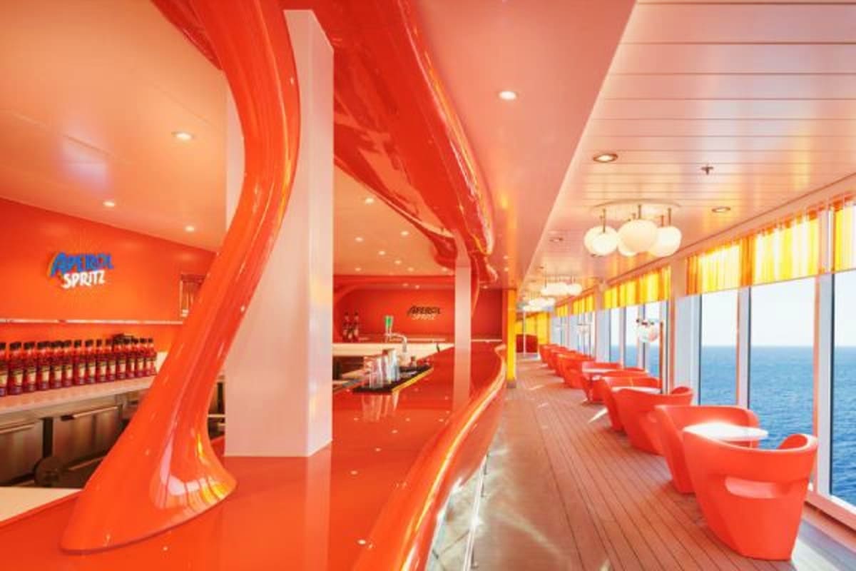Modern red and orange interior of a cruise ship bar with seating areas, ocean views through large windows, and contemporary d