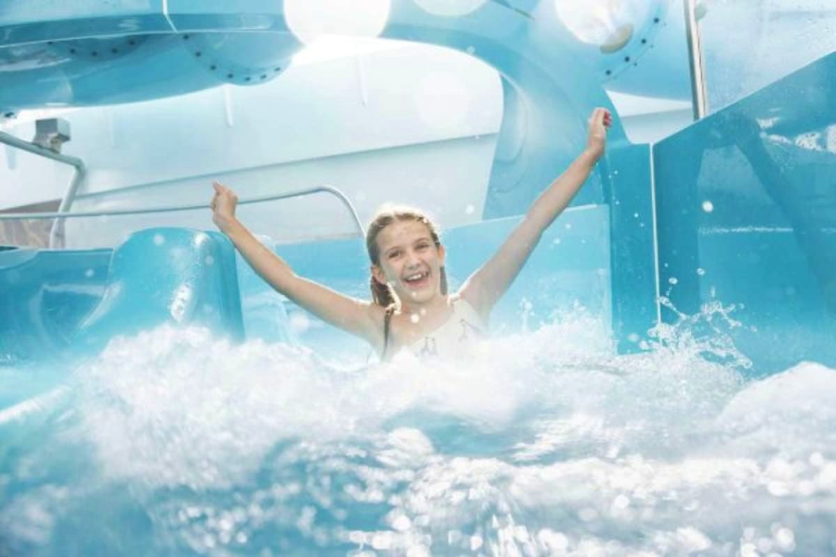 Child enjoying water slide at cruise ship pool with arms raised in excitement and splashing water