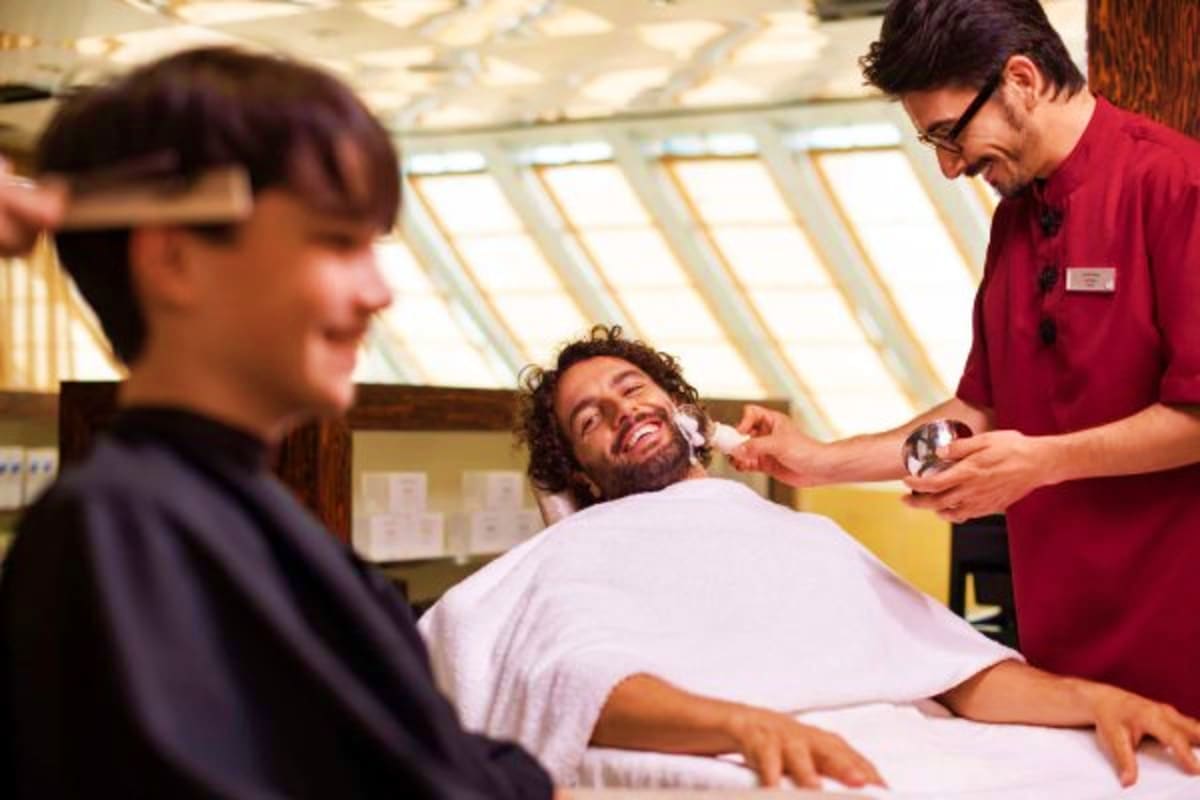 Barber styling customer's hair and beard inside a cruise ship salon with skylights overhead