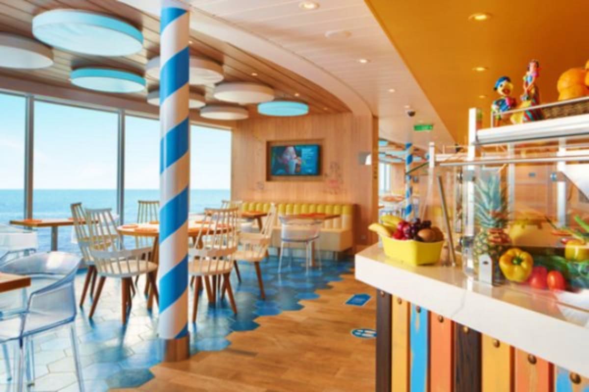 Modern cruise ship dining area with ocean view windows, blue and white striped column, yellow ceiling, and fresh produce on c