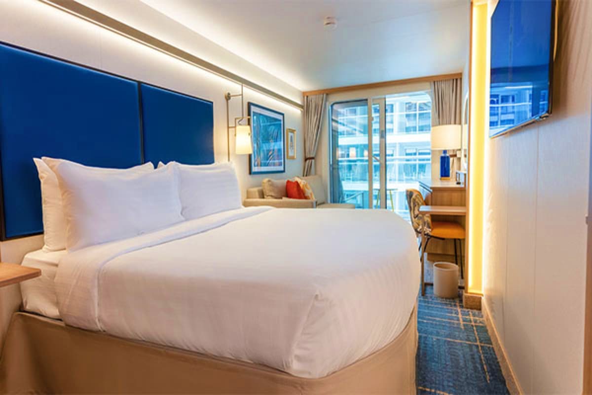 Modern cruise ship stateroom with queen bed, blue headboard, floor-to-ceiling windows overlooking ocean and city skyline