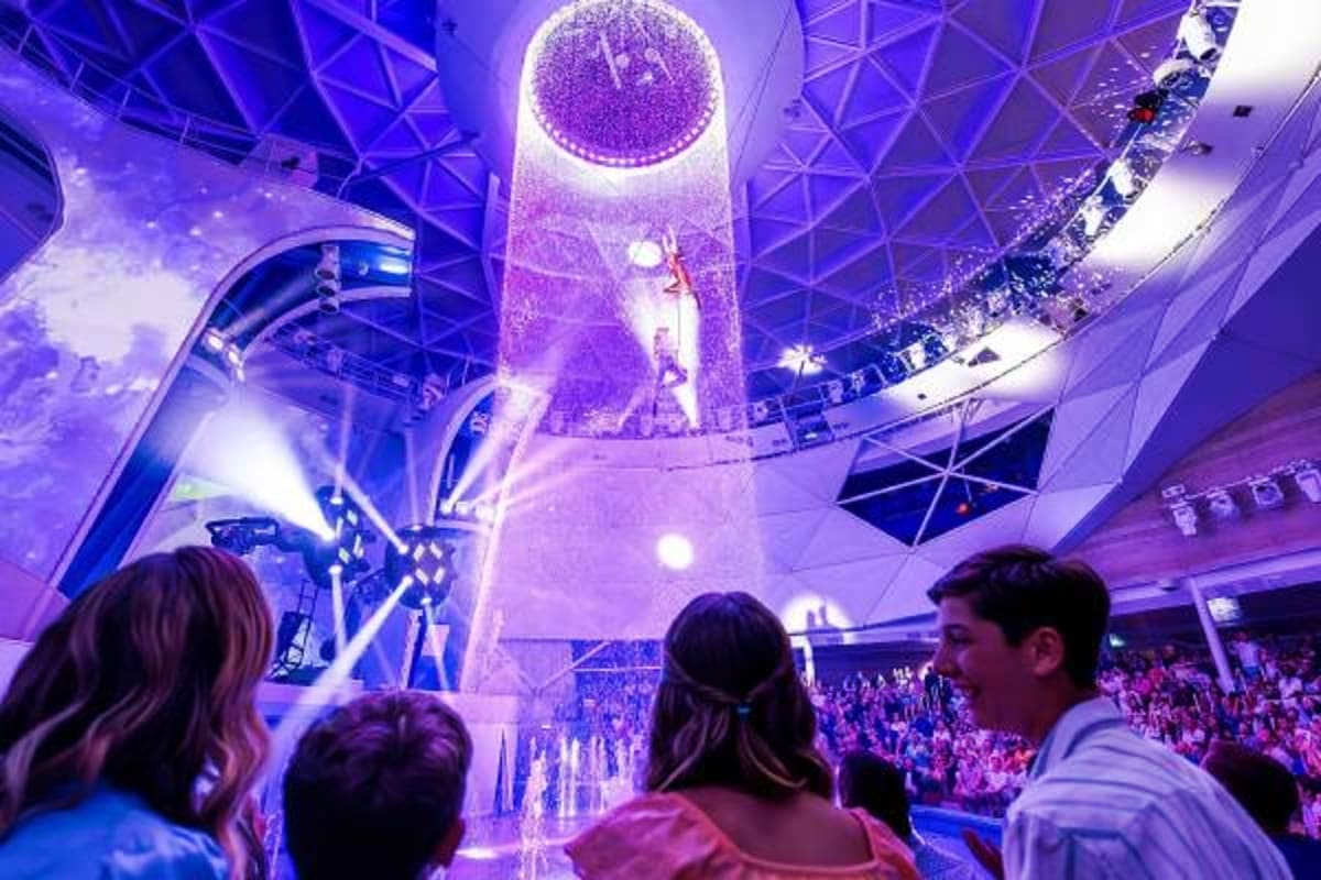 Audience watching aerial acrobat performance in colorful purple-lit theater atrium on cruise ship with multiple deck levels v