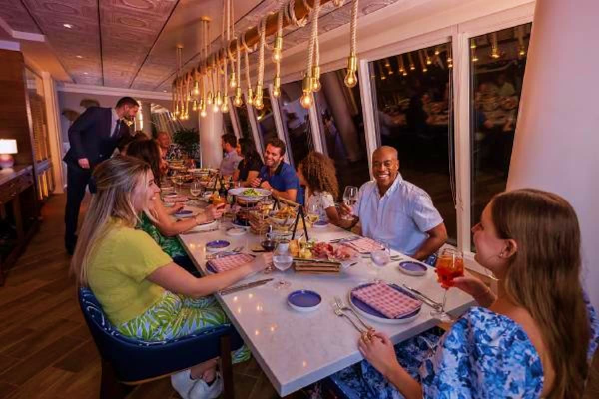 Guests dining together at an elegant indoor restaurant on a cruise ship with pendant lighting and ocean views through windows