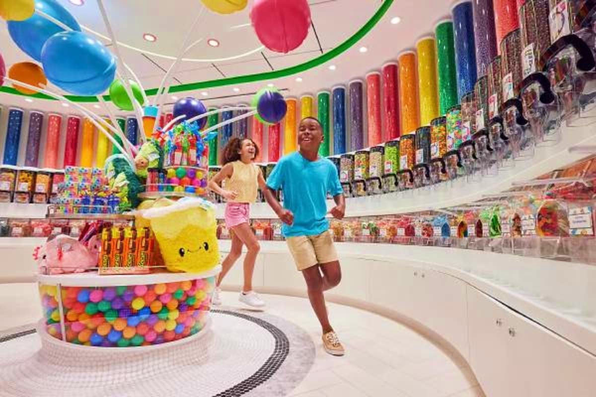 Children playing in a colorful toy store with rainbow-colored shelves, hanging balloons, and a ball pit display on a cruise s