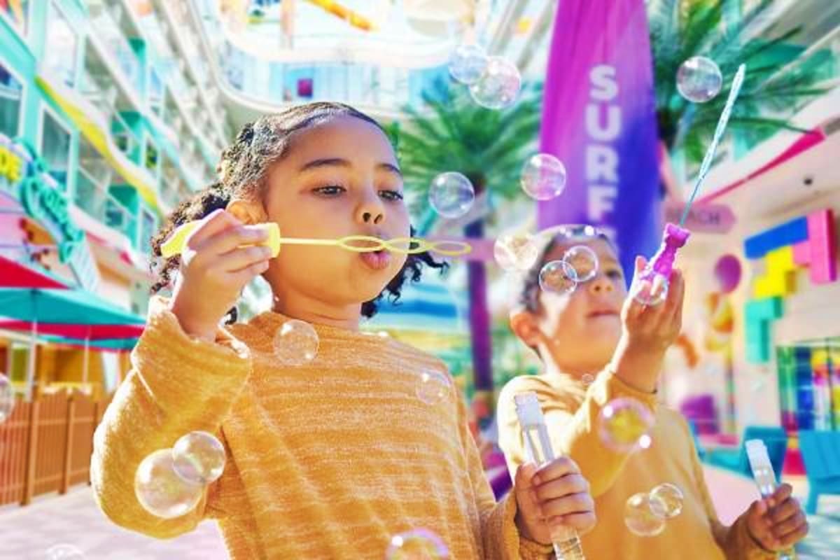 Children blowing bubbles at a colorful indoor atrium on a cruise ship with tropical plants and bright signage in background