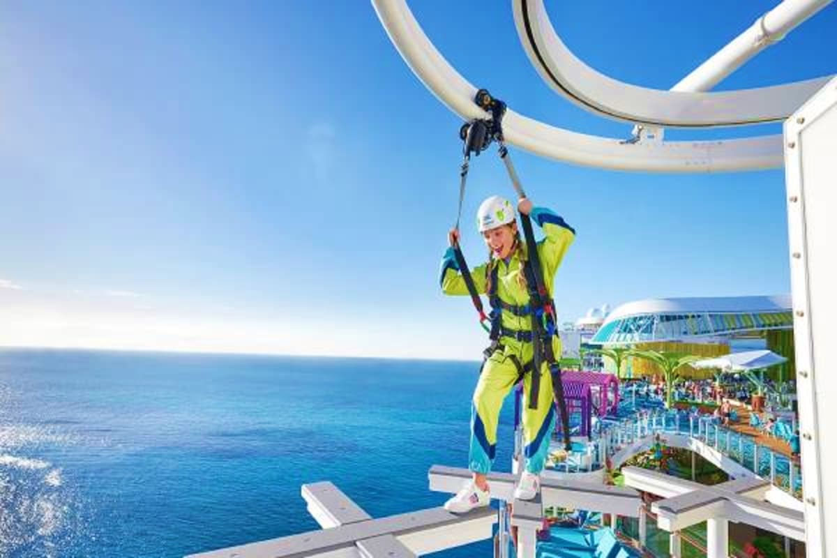 Person in yellow safety gear zip-lining above ocean from cruise ship deck with blue water and ship structures visible below.