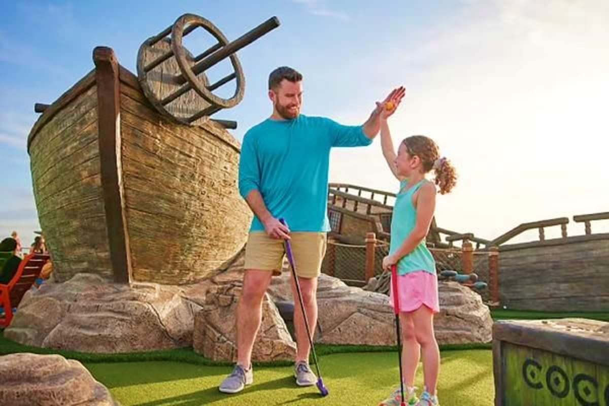 Father and daughter playing mini golf at cruise ship port with wooden pirate ship and steering wheel decoration