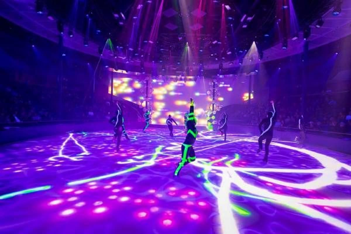Neon-lit nightclub interior with purple and green LED dance floor, performers in glowing outfits, and illuminated ceiling pan