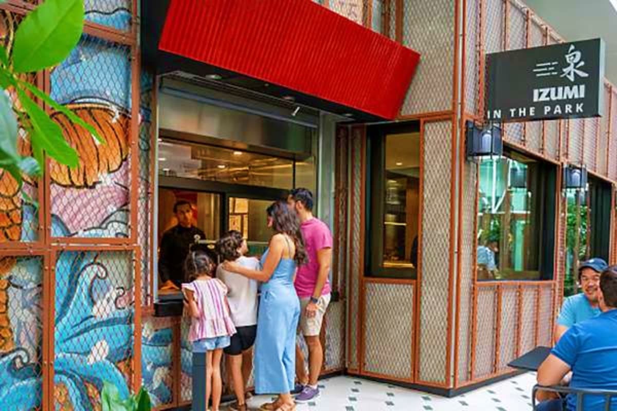 Customers queuing outside Izumi In The Park restaurant storefront with red awning and decorative tropical artwork on exterior