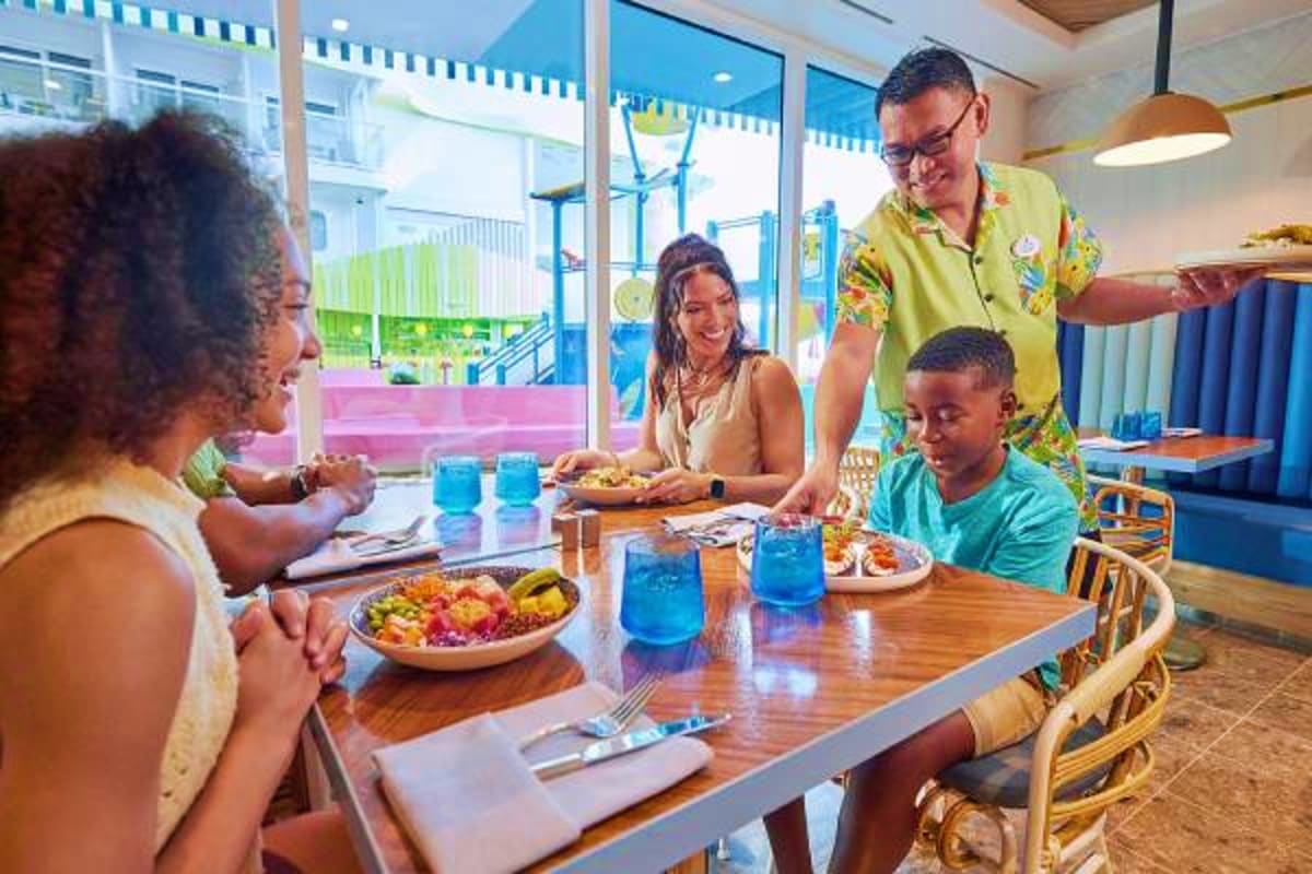 Family dining together at a wooden table in a bright cruise ship dining room with large windows overlooking the ocean and shi