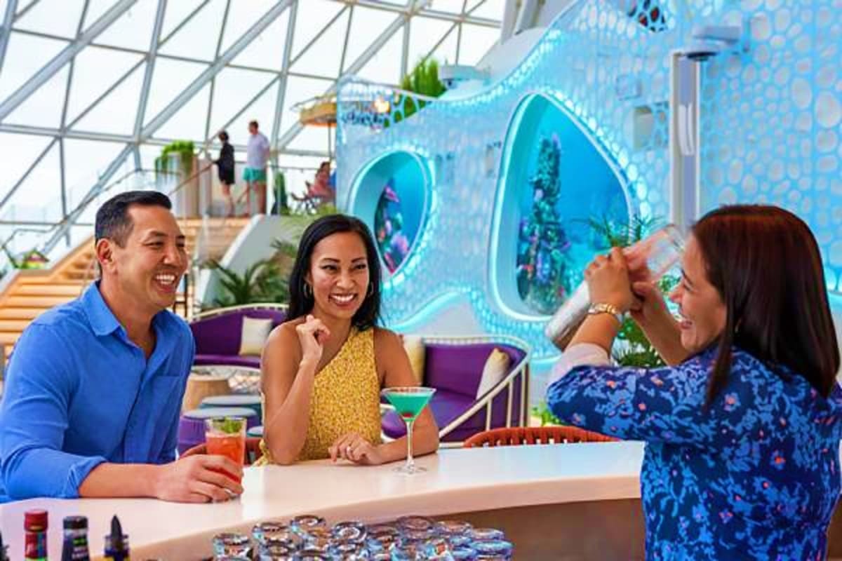Three guests enjoying cocktails at an indoor bar with blue aquarium-themed wall panels and glass ceiling in a cruise ship atr