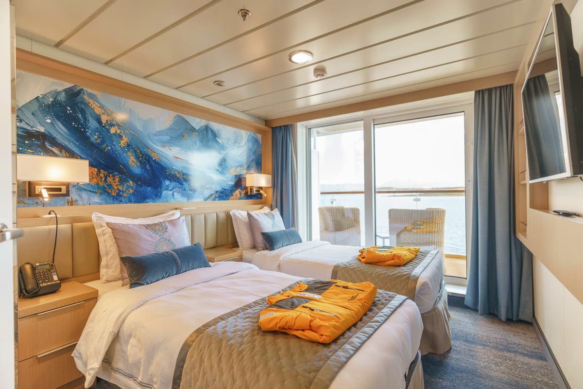 Arctic Wonders of Greenland and Iceland Modern cruise ship cabin with two beds, mountain landscape artwork, ocean view balcony, and arctic expedition jackets