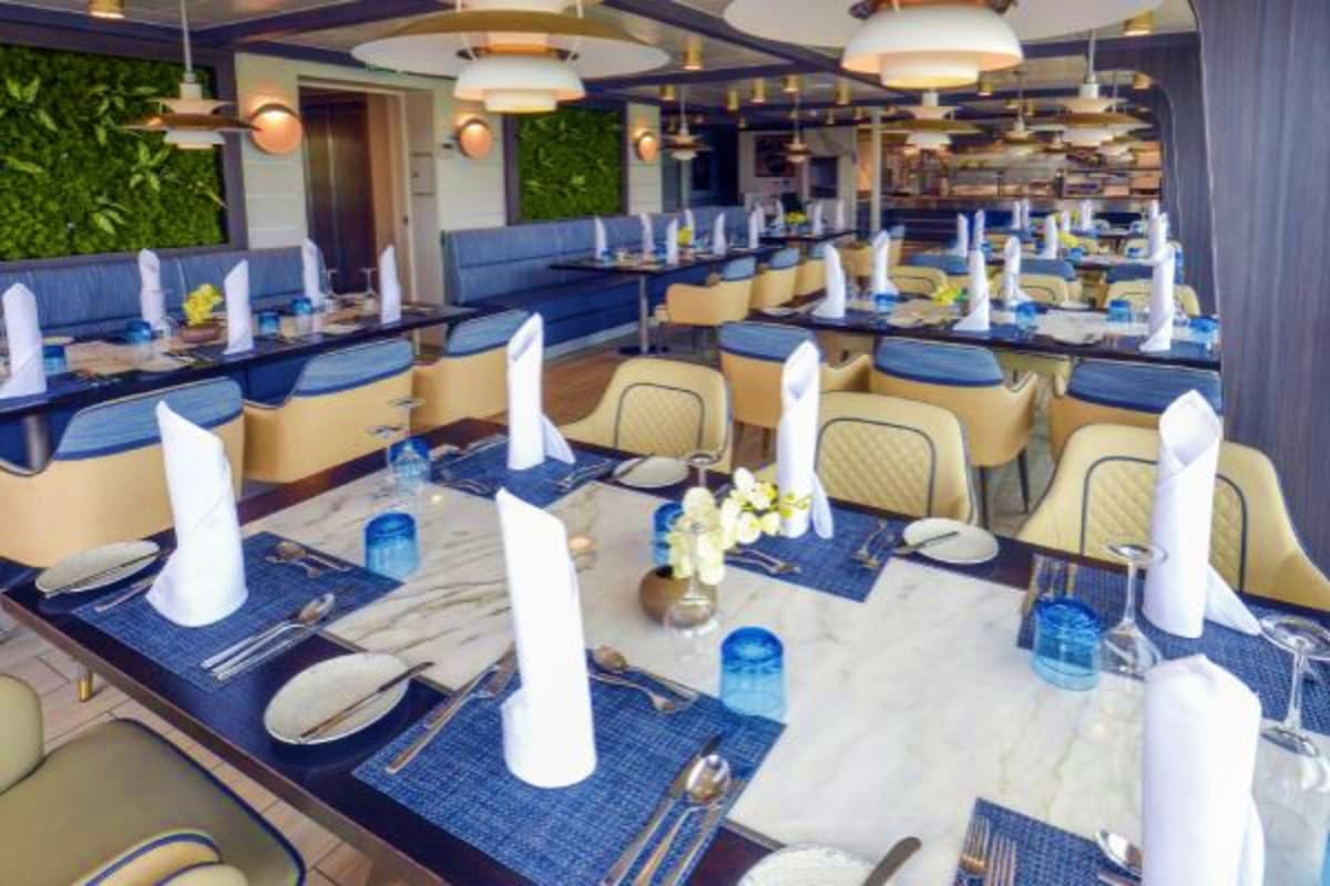 Arctic Wonders of Greenland and Iceland Modern cruise ship dining room with blue and cream upholstered banquette seating, white napkins, marble tables, and pendant l