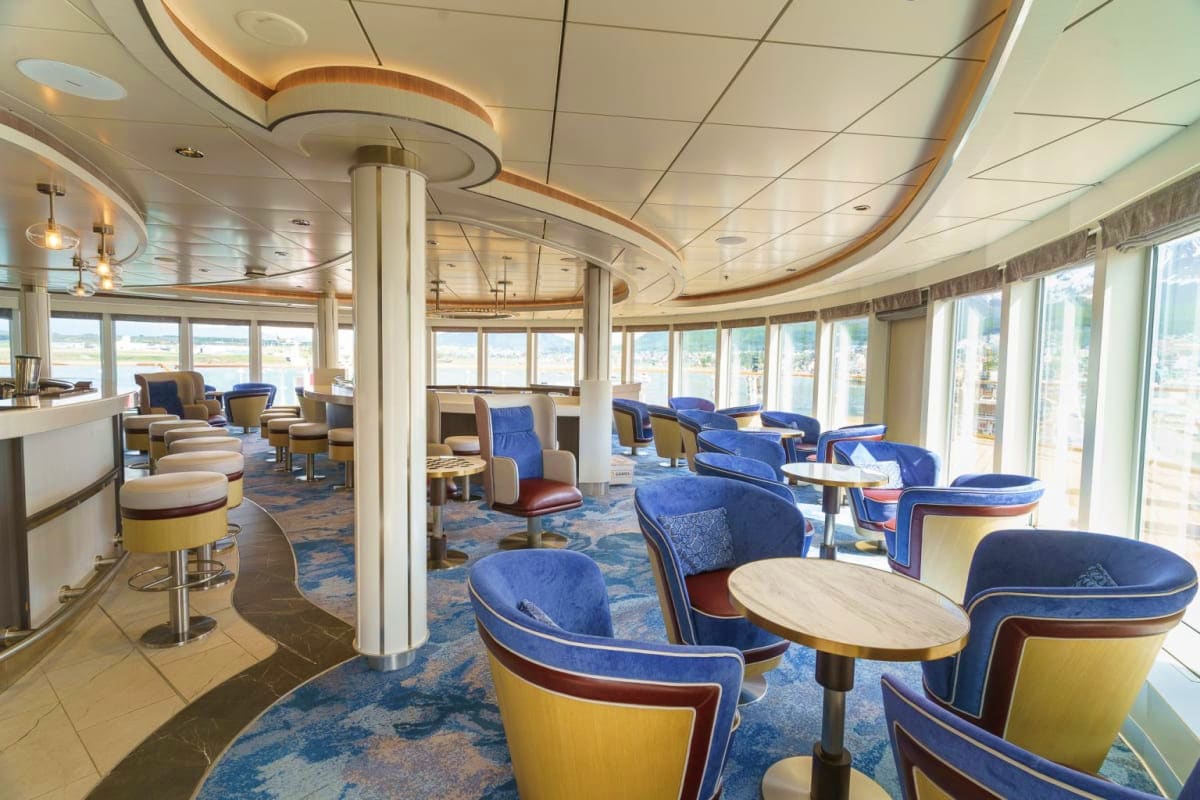 Modern cruise ship lounge with curved ceiling design, blue and yellow seating, tables, and panoramic windows overlooking ocea