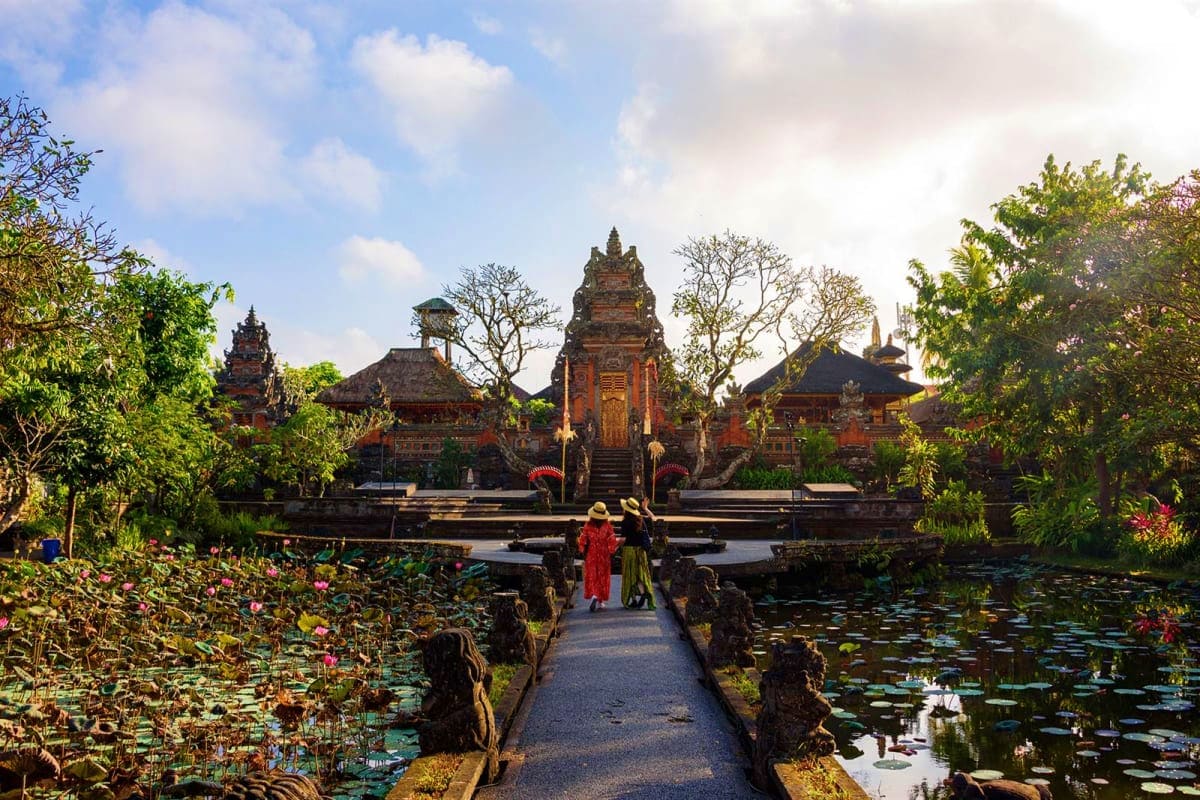Balinese temple with red brick architecture reflected in lily pond, flanked by lush tropical gardens and stone pathway