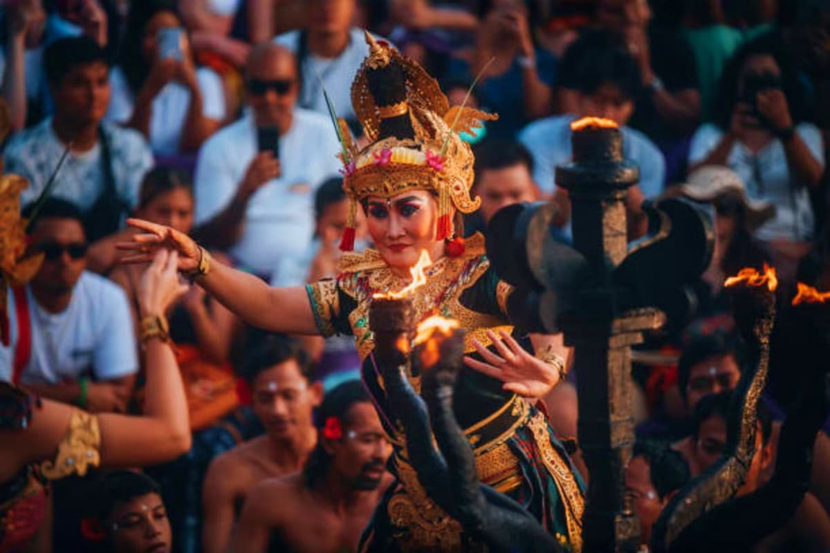 Performer in ornate gold costume and headdress dancing at Balinese cultural festival with fire performers and crowd in backgr