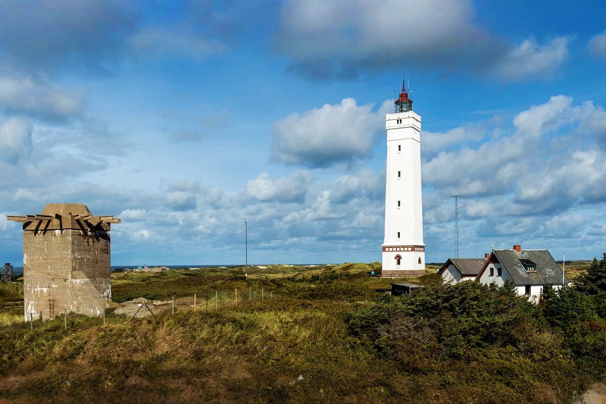 From Frankfurt to Copenhagen White lighthouse with red top on coastal dunes next to keeper's house and ruins of old windmill under blue sky with clouds