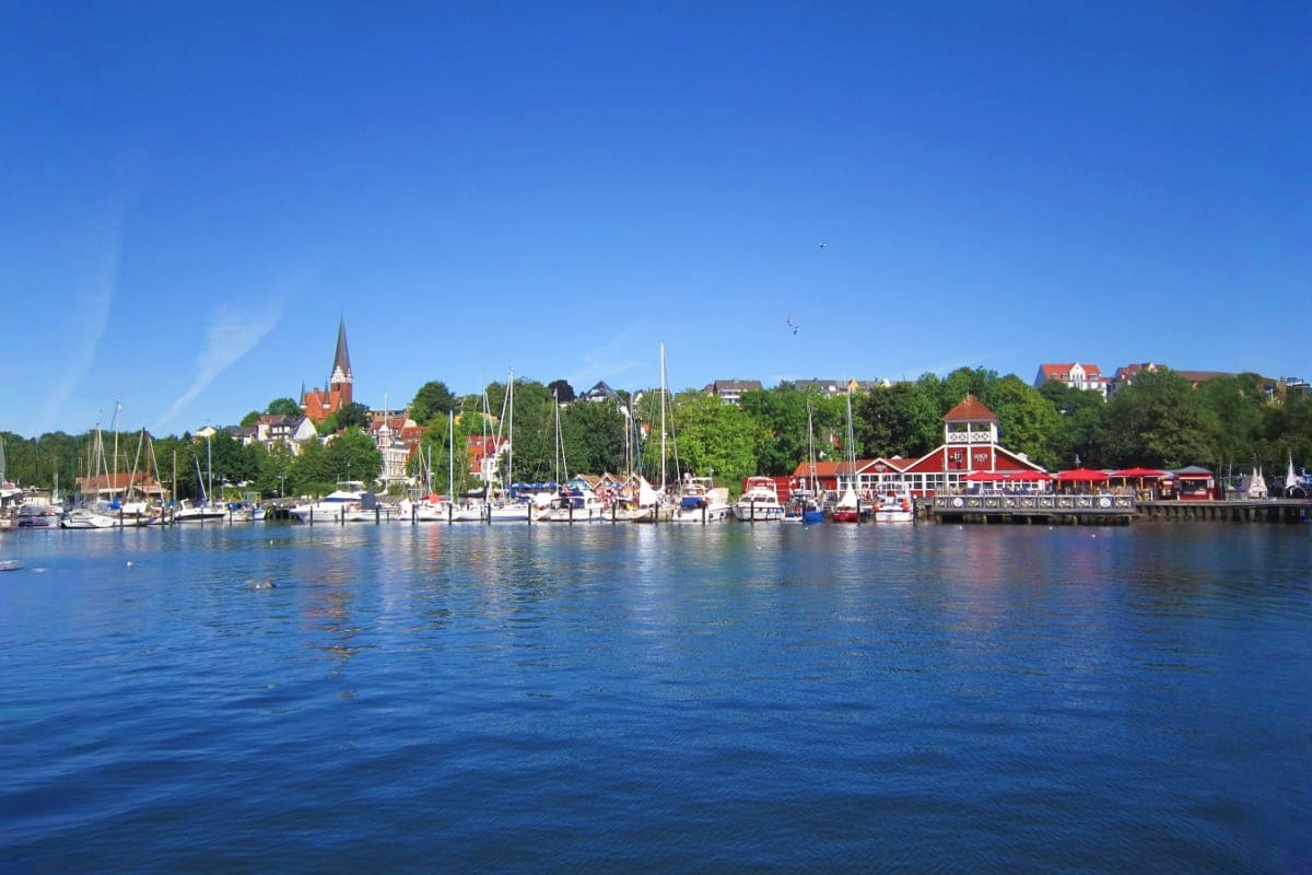 From Frankfurt to Berlin Scenic waterfront harbor view with sailboats, red Victorian buildings, church spire, and tree-covered hills under clear blue