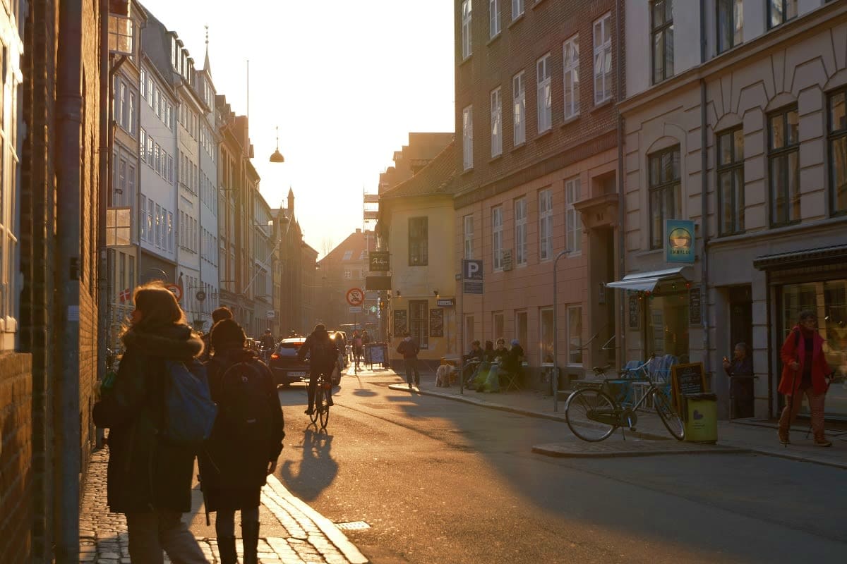 Pedestrians and cyclists on a narrow European cobblestone street lined with historic brick buildings at golden hour sunset