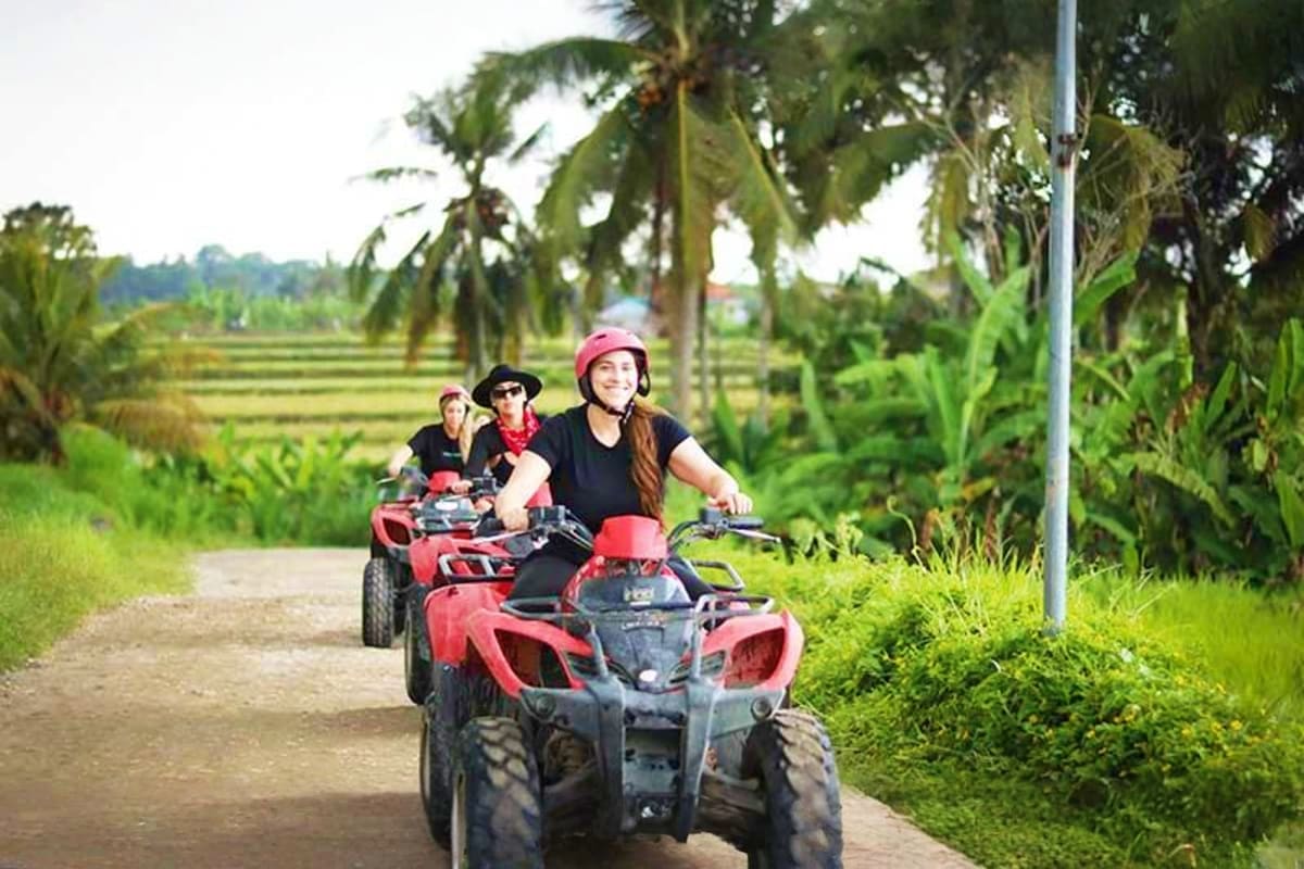 Three people riding red and black ATVs on a rural dirt road lined with palm trees and rice paddies in Bali