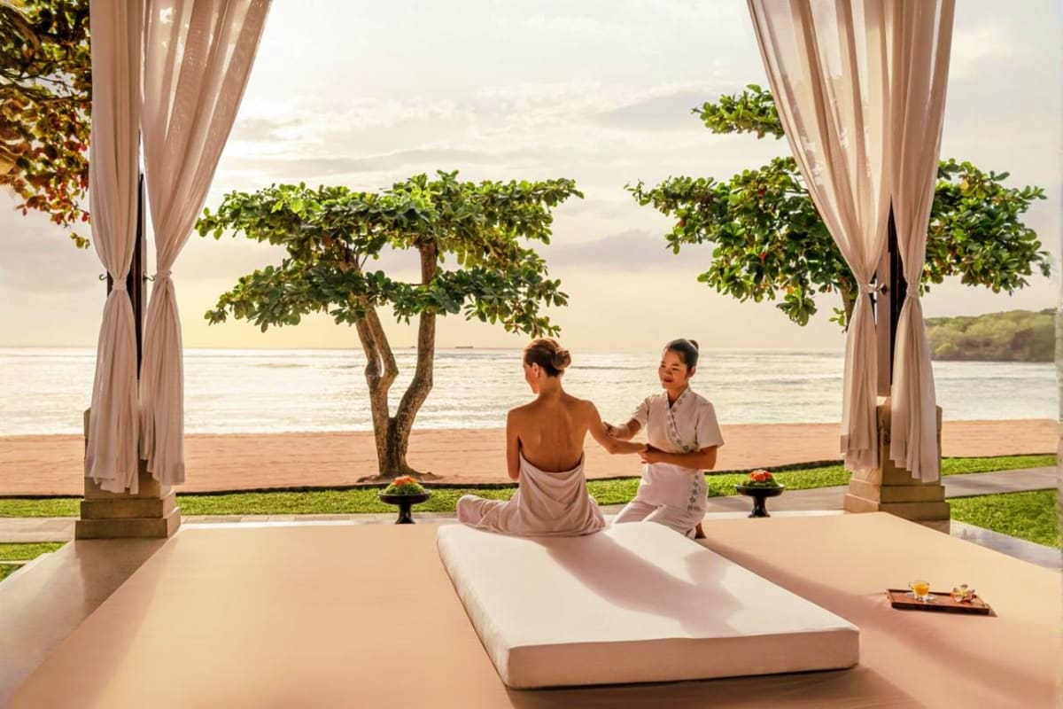 Woman receiving massage treatment in beachfront spa pavilion overlooking ocean and tropical tree in Bali