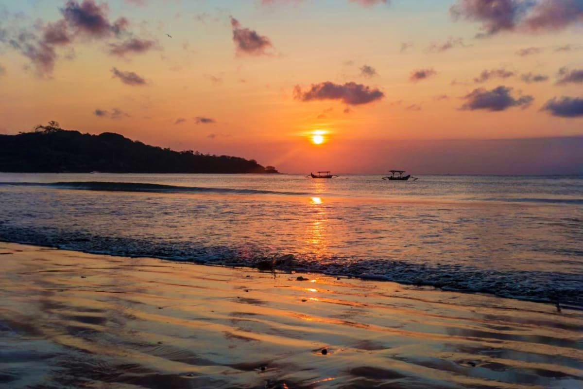 Sunset over Bali beach with traditional fishing boats, golden sky reflecting on wet sand and calm ocean waters