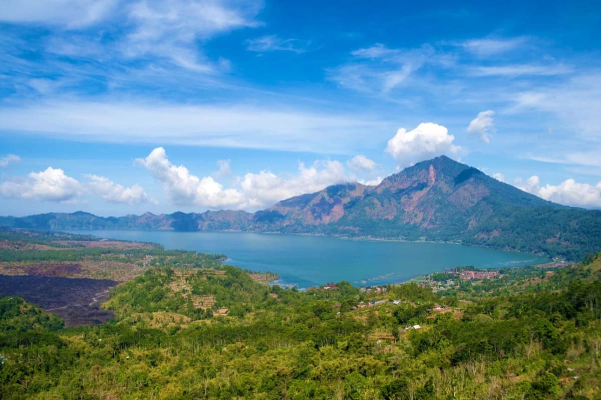 Bali Nature and Luxury Package Aerial view of Mount Batur volcano overlooking turquoise lake with lush green forests and village settlements in Bali, Indone