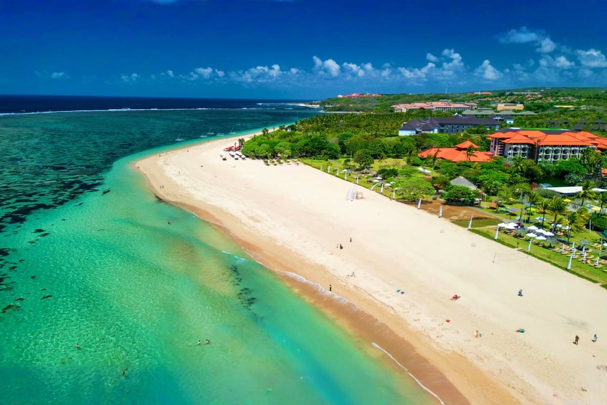 Aerial view of a pristine white sand beach with turquoise water, luxury resort with orange roofs, and lush tropical vegetatio