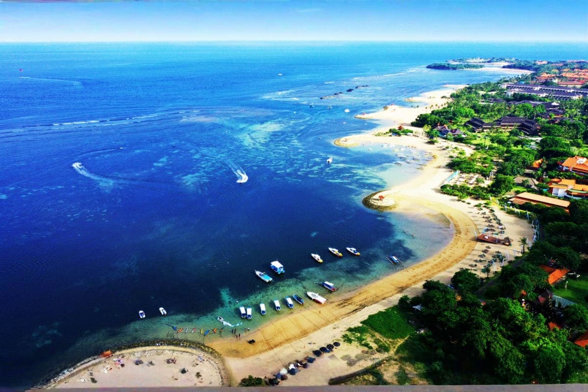 Aerial view of Bali beach resort with turquoise waters, white sand, moored boats, tropical vegetation, and coastal villa deve
