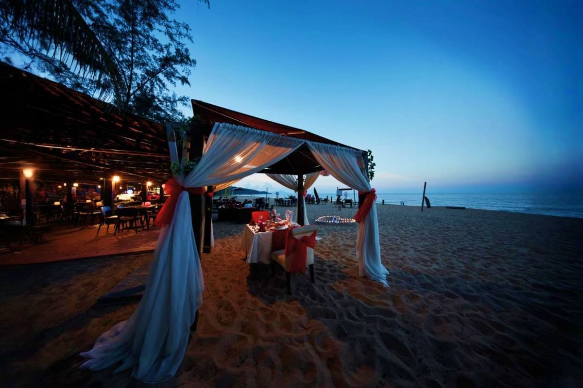 Beachfront dinner setup at dusk with white draped canopy tent, red chairs, and decorated table overlooking calm sea under cle