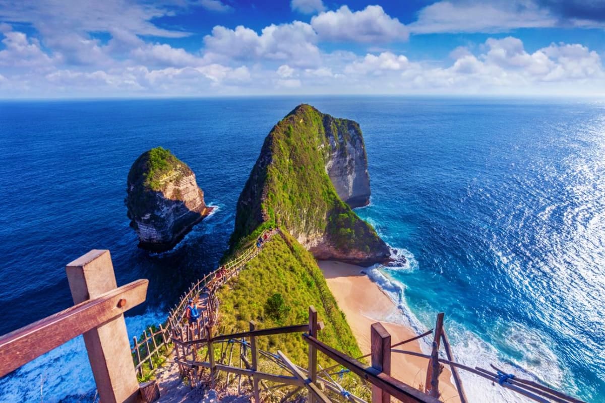 Wooden staircase descending a lush green cliff overlooking Kelingking Beach in Nusa Tenimber, Bali, with turquoise ocean and 