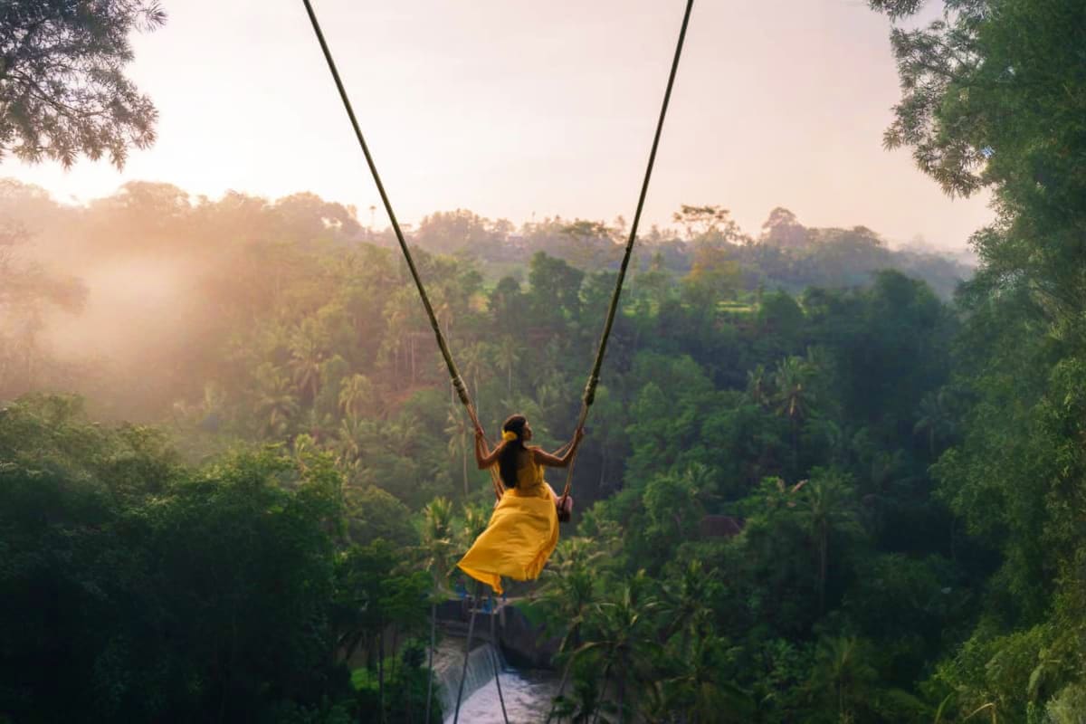 Woman on jungle swing wearing yellow shirt overlooking misty rainforest valley at sunrise in Bali