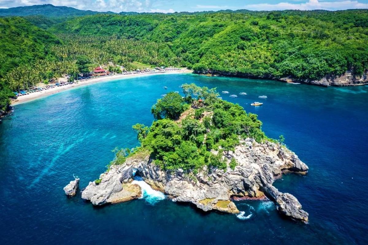 Aerial view of a rocky island with lush green vegetation in turquoise blue waters, with a sandy beach and forested coastline 