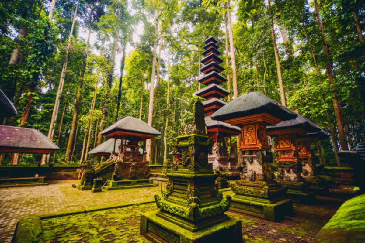 Ancient Hindu temple with multi-tiered pagoda surrounded by tall trees and moss-covered stone structures in Bali forest sanct
