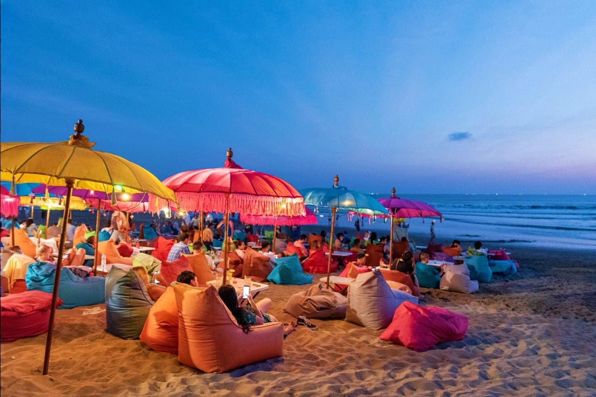 Colorful beach lounge area at sunset with bean bag seating, decorative umbrellas, and beachgoers enjoying the ocean view in B