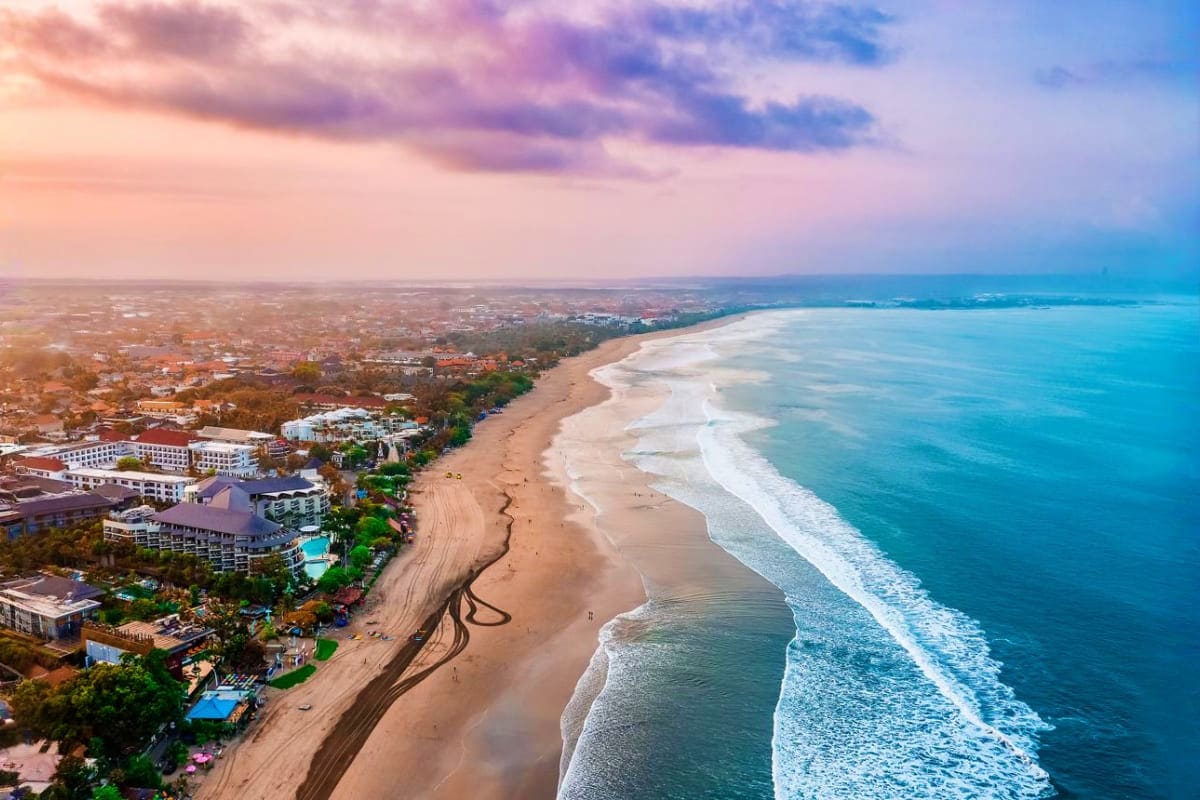 Aerial view of a coastal town with sandy beach, resort buildings, and turquoise ocean waves at sunset in Bali