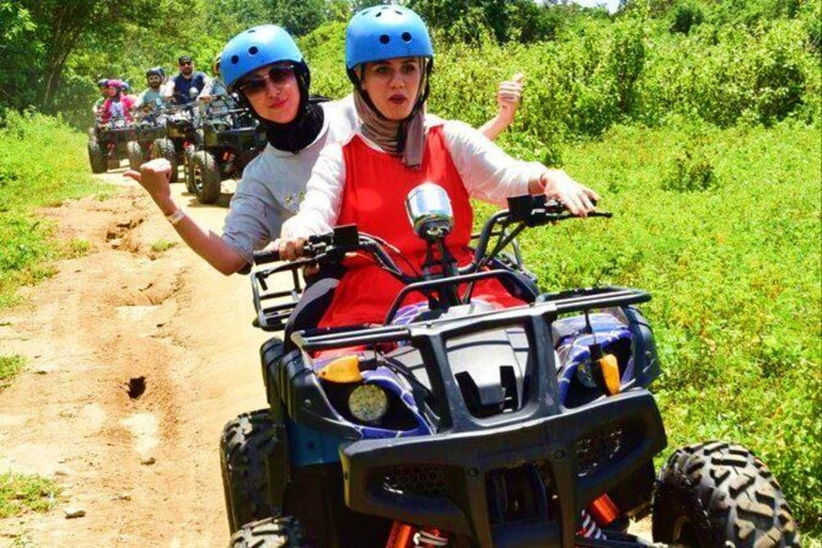 Two women wearing blue helmets riding ATVs on a dirt trail through lush green forest with other riders visible behind them