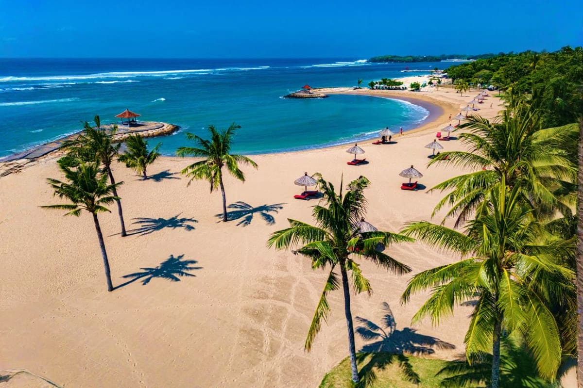 Arabian Honeymoon Escape to Indonesia Aerial view of a tropical beach in Indonesia with turquoise waters, white sand, palm trees, and beachgoers under umbrellas