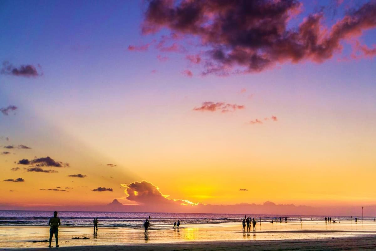Arabian Honeymoon Escape to Indonesia Beachgoers and swimmers on a sandy beach at golden hour sunset with purple and orange sky over the ocean