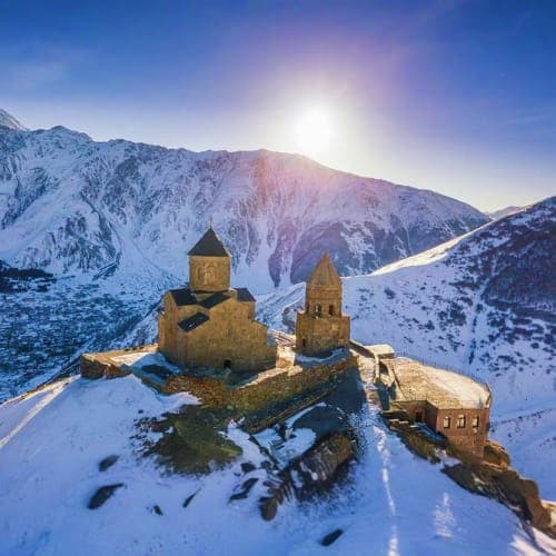 Ancient stone churches with golden domes on snow-covered island surrounded by frozen lake and snow-capped Caucasus mountains 