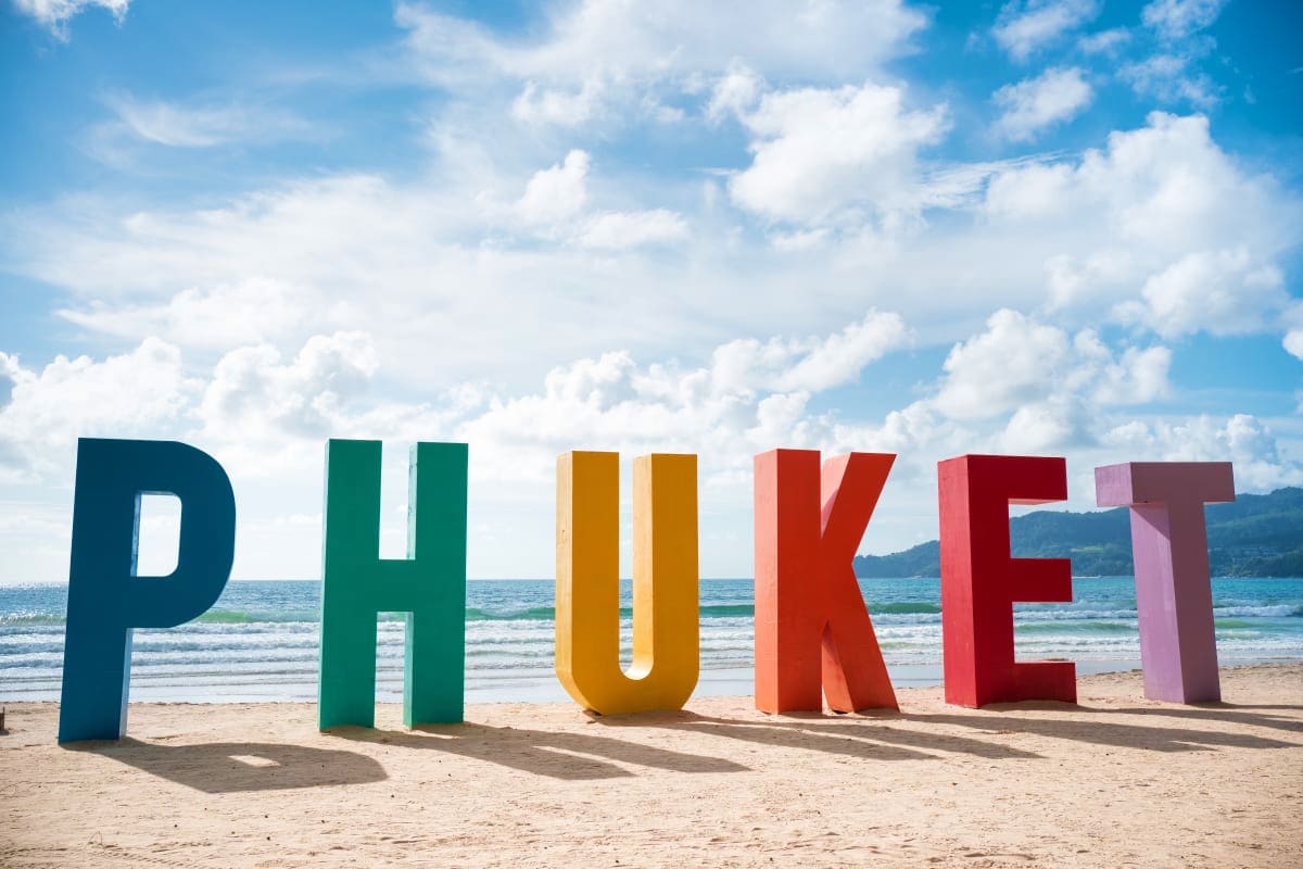 Colorful Phuket sign on sandy beach with ocean waves and mountains in background under blue sky