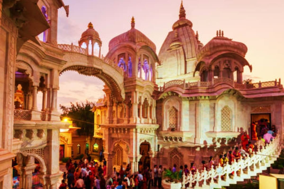 Illuminated Hindu temple with ornate domes and spires at dusk, crowds of visitors gathering in courtyard during spiritual pil