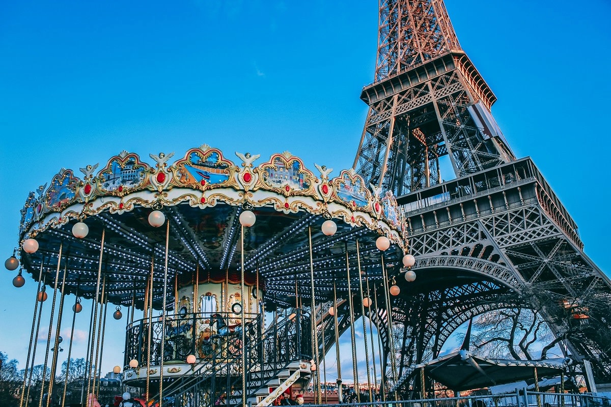 Vintage carousel with ornate golden crown and hanging lights beneath the Eiffel Tower in Paris on clear blue sky day