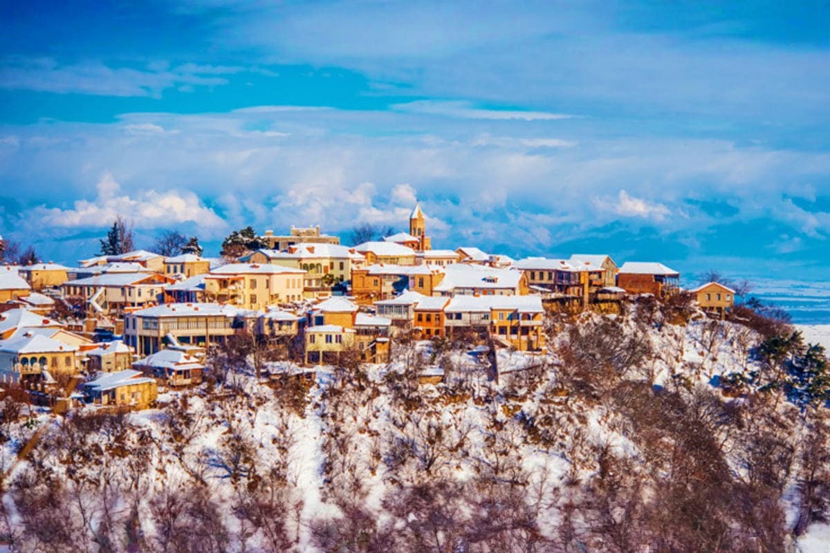 Snow-covered hillside village with yellow and orange buildings and a church steeple against blue sky and distant mountains