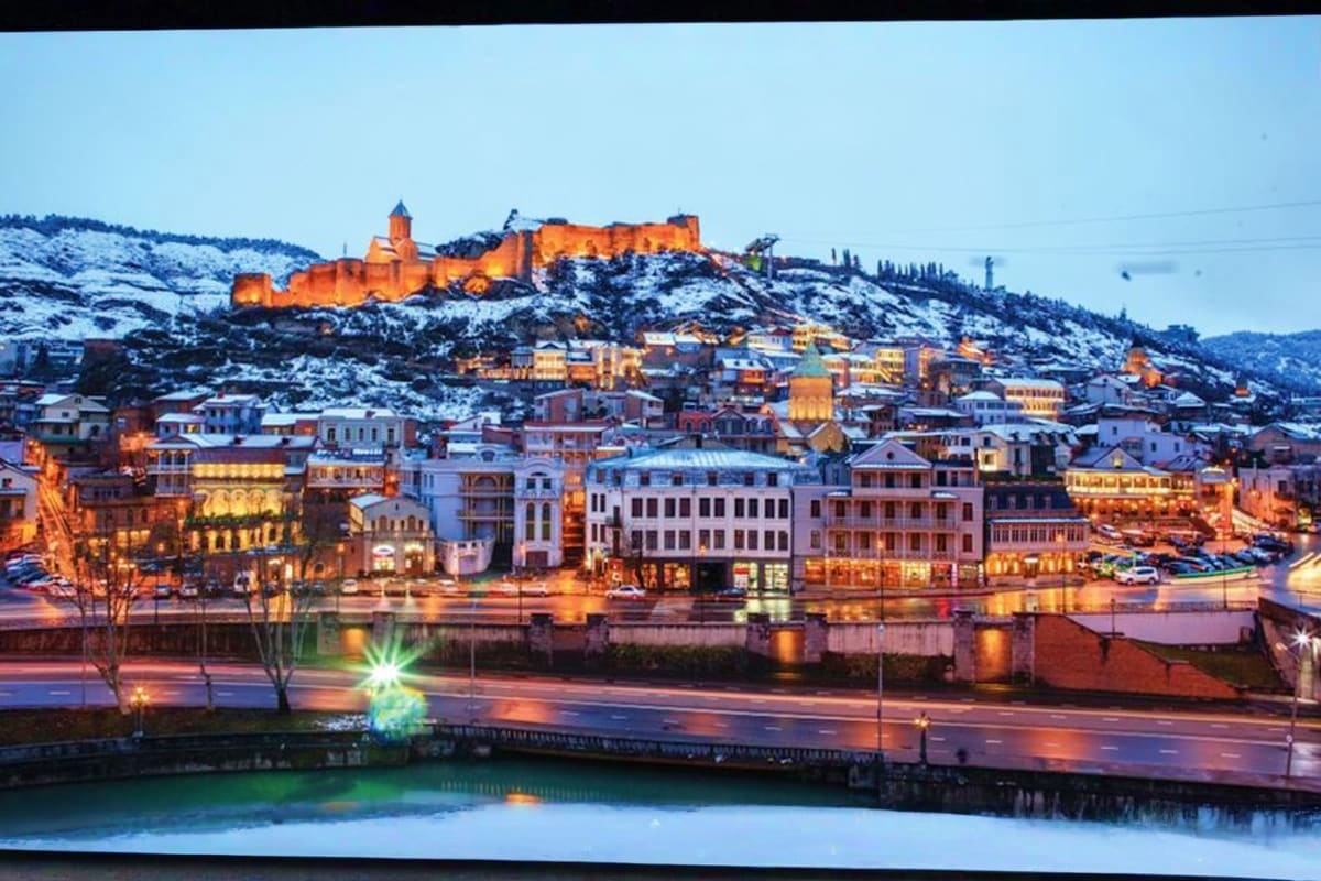 Tbilisi cityscape at dusk with illuminated Narikala Fortress on snowy hilltop, historic buildings reflected in Mtkvari River 