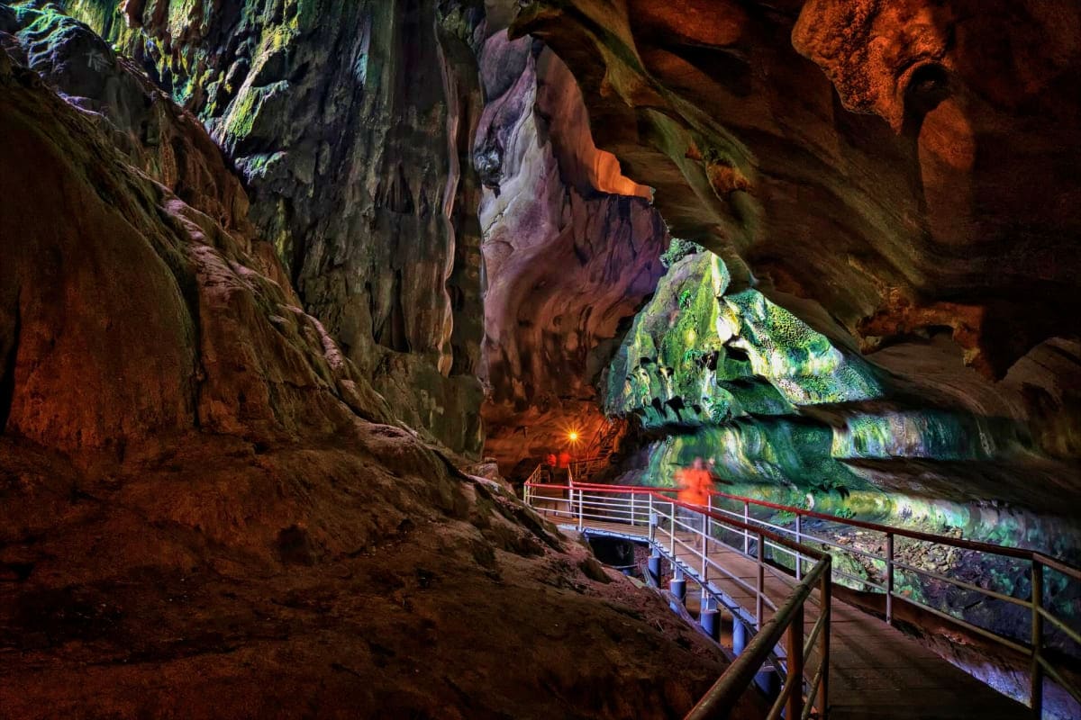 Underground cave chamber with moss-covered rock formations, illuminated walkway with metal railings, and underground lake or 