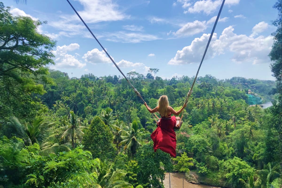 Woman in red dress on jungle swing overlooking lush forest, river, and palm trees in Bali, Indonesia