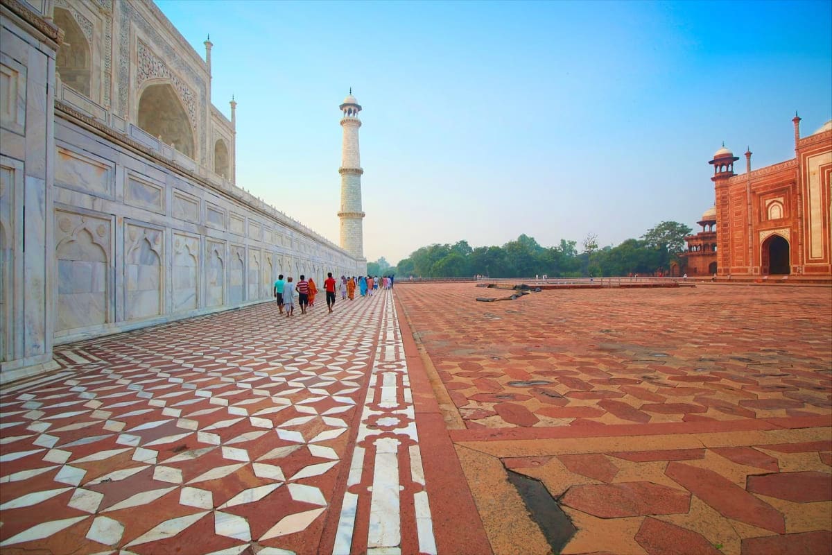 Taj Mahal courtyard with geometric red and white marble floor, minaret in background, and visitors walking along the mosque w
